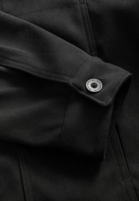 Nærbillede af sort jakke stof, med en knap med præget tekst, fin tekstur og synligt syning på kraven og ærmet.