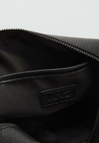 Sac noir à fermeture éclair avec un extérieur texturé, doté d'un intérieur en tissu lisse et d'un petit patch de marque. Design principalement monochrome.