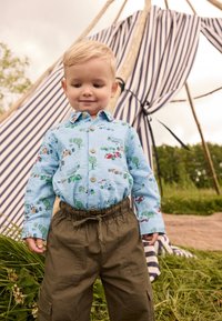 Camicia azzurro chiaro con bottoni e stampa di veicoli colorati; pantaloni cargo verde oliva con vita elastica; all'aperto con una tenda a righe sullo sfondo.