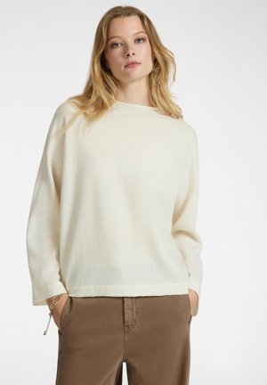 Vrouw met blond haar in een crèmekleurige, losse trui en een paarse broek, staande met haar handen in haar zakken tegen een effen achtergrond.