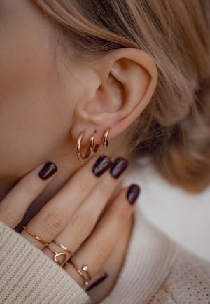 Goldene Creolen in drei Größen hängen vom Ohr, ergänzt durch goldene Ringe an den Fingern und dunklen Nagellack. Leichter, strukturierter Pullover.