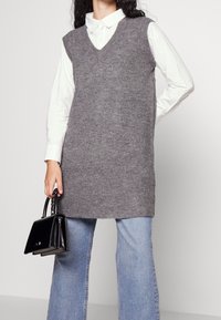 Henkilö, joka pitää yllään harmaata ilman hihoja neuleliiviä valkoisen nappipaidan päällä, vaaleansiniset farkut jalassa ja kädessään pieni musta käsilaukku.