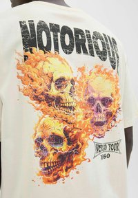 Krémové tričko s grafikou troch plameňom pokrytých lebiek s prasklinami. Nápis "NOTORIOUS" je vytlačený nad nimi tučnými čiernymi písmenami.