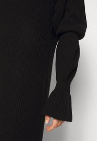 Svart ribbad stickad klänning med långa ärmar och utsvängda mudd avslut samt en texturerad yta. Tyget verkar mjukt och tjockt.