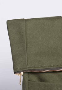 Bolsa de lona verde oliva con un diseño de parte superior enrollable y una cremallera negra con herrajes dorados en la apertura, que muestra una superficie texturizada.