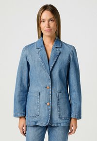 Sininen farkkutakki, jossa on leikattu kaulus, kaksi etutaskua ja ruskeat napit. Takki on rento malli, jossa on näkyvät ompeluyksityiskohdat.