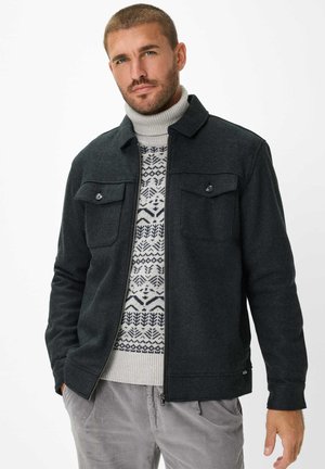 Dunkelgraue Jacke mit Kragen, Frontreißverschluss und zwei Brusttaschen, darüber ein helles, geometrisch gemustertes Sweatshirt.