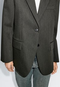Donkergrijze blazer van gestructureerde stof, met een sluiting met twee knopen, notch-lapels en een enkele borstzak, gedragen met spijkerbroeken.
