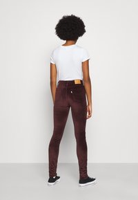 Leggings de pana en color burdeos, con un diseño ajustado, textura suave, cintura alta con una etiqueta marrón clara en la parte trasera, combinados con una camiseta blanca y zapatillas negras.