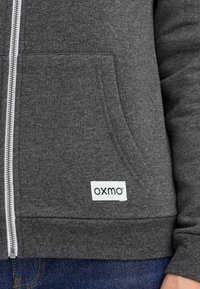 Grijze rits hoodie van zachte stof, met een voorzak, geribbelde boorden en een "OXMO" label aan de onderkant aan de linkerkant.