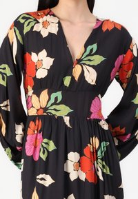 Svart blommig klänning med långa ärmar, som har livfulla röda, rosa och gröna blommor, en djup V-ringning och en rynkad midja.