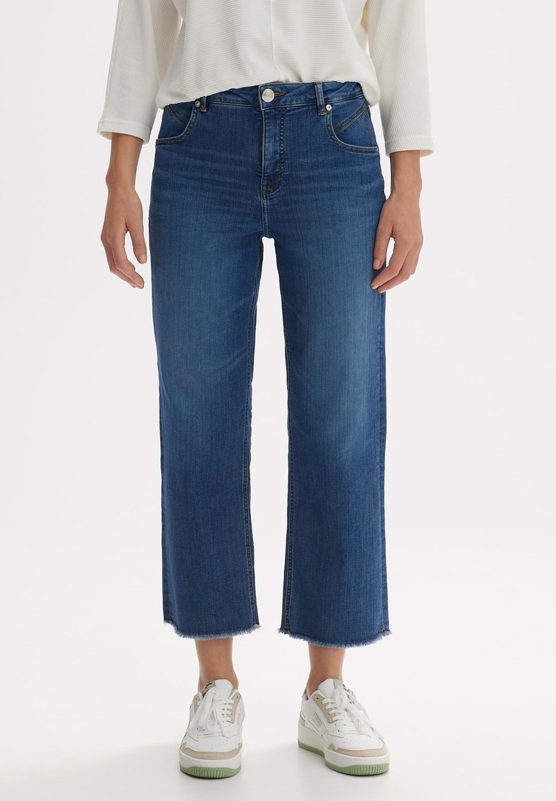 Personne portant un jean bleu court à jambes larges avec des ourlets bruts, un haut blanc à manches longues et des baskets blanches avec des accents beiges.