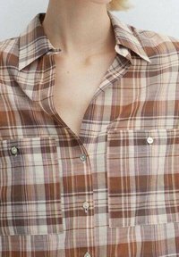 Donna che indossa una camicia a quadri marrone, beige e rosa con due taschini sul petto e colletto aperto che mostra il collo e la parte superiore del petto.
