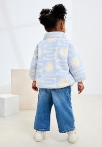 Fleece jacket in light blue and cream with sun patterns, paired with wide-leg blue denim pants and white sneakers.