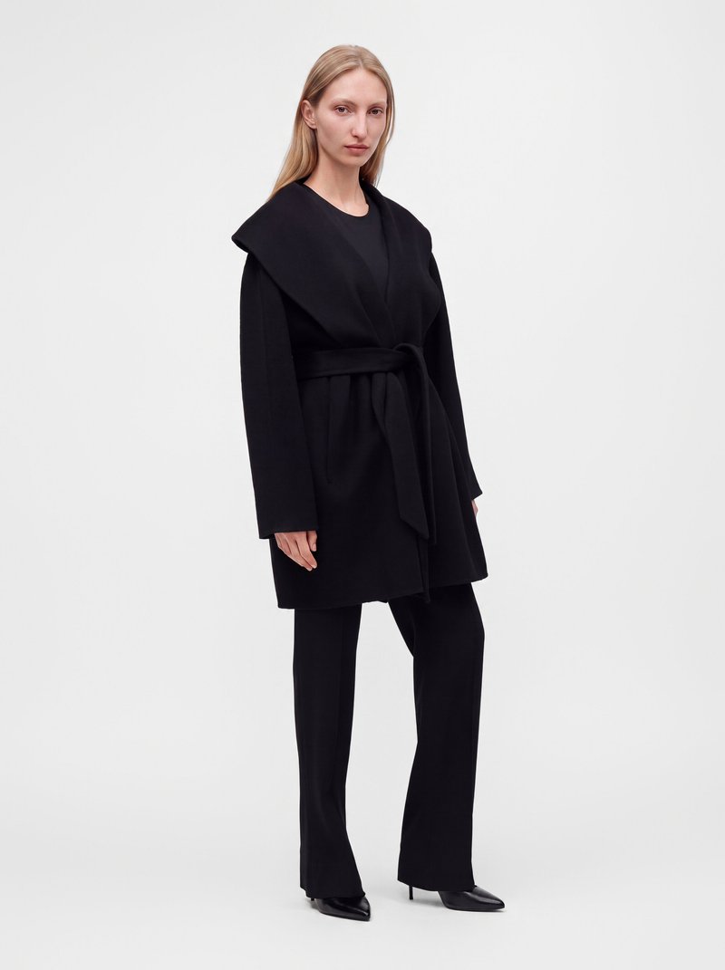 Femme aux longs cheveux blonds portant un manteau noir ceinturé, un pantalon noir et des escarpins noirs à bout pointu, debout devant un fond clair uni.