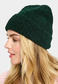 Dark green knitted beanie with a textured fabric, tapered design, and ribbed edge, worn on a model with long blonde hair.