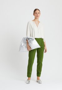 Weißes Blusenshirt mit Schleifendetail, kombiniert mit grünen, taillierten Hosen und silbernen Slippern, hält eine zerknitterte silberne Tasche. Minimalistisches Design.