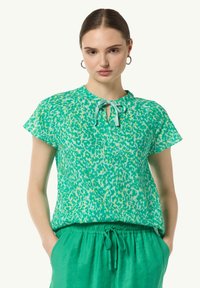 Green short-sleeve top with a leopard print pattern, featuring a keyhole neckline tied with a ribbon. Lightweight, smooth fabric.