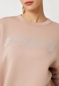 Felpa rosa con la scritta "GUESS" in strass argentati, con scollatura rotonda e texture morbida e liscia. Design casual adatto per l'uso quotidiano.