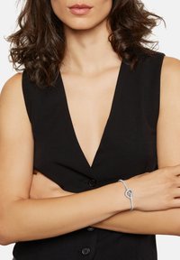 Bracciale in metallo grigio con un design a forma di cuore, indossato su un polso. Sullo sfondo si intravede un abbigliamento senza maniche di colore nero con bottoni.
