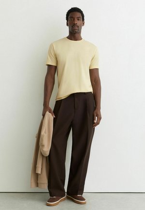 Mand står mod en hvid væg iført lysegul t-shirt, mørkebrune bukser med vide ben og brune sko, holder en beige sweatshirt i venstre hånd.