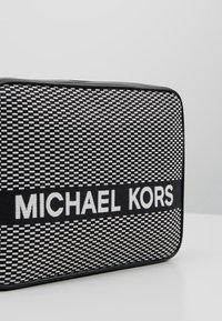 Svart och vit Michael Kors-väska med ett geometriskt vävt mönster, med en framträdande logotyp i vitt och en elegant rektangulär form.