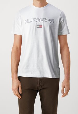 Mann trägt ein weißes Kurzarm-T-Shirt mit dem Schriftzug "HILFIGER '85" und einem kleinen rechteckigen Flaggenlogo, kombiniert mit dunkelbraunen Hosen.