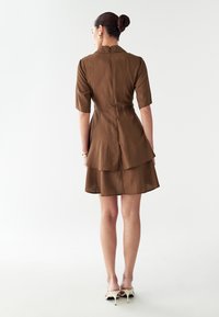 Rjava večplastna obleka s kratkimi rokavi, teksturirana tkanina, zadrga na hrbtu in prilegajoč korzet. Nošena z svetlimi škornji.