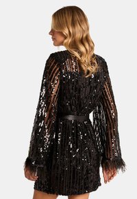 Abito nero in paillettes con maniche lunghe e trasparenti, vita sfiancata e dettagli in piume ai polsini. Presenta una linea dritta e una texture lucida.