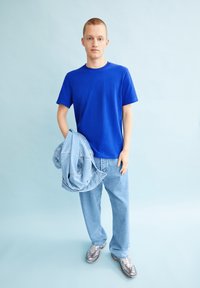 Blaues Baumwoll-T-Shirt mit Rundhalsausschnitt, kombiniert mit hellblauen Jeans. Die Person hält eine Jeansjacke. Die Sneakers haben ein gemustertes Design.