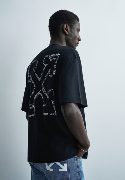 Homme portant un t-shirt graphique noir avec un design abstrait de texte blanc et un jean bleu avec imprimé flèche blanche, debout devant un fond gris clair.