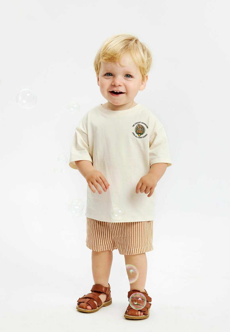 Krämfärgad t-shirt med kort ärm och rund grafik, kombinerad med bruna och vita randiga shorts och bruna sandaler. Bubbles är i närheten.