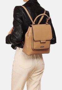 Zaino in pelle tan con un design strutturato, chiusura a ribalta e cuciture nere a contrasto. Spallacci regolabili e maniglia superiore piccola.