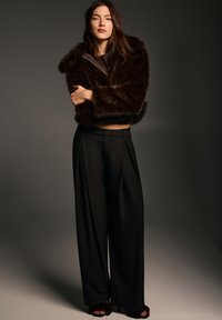 Giacca corta in pelliccia sintetica marrone, pantaloni neri larghi con pieghe frontali e calzature marroni aperte, su uno sfondo scuro.