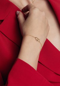 Złota bransoletka w kształcie serca z cienkim łańcuszkiem, noszona na dłoni na tle czerwonego materiału. Prosty design z polerowanym wykończeniem.