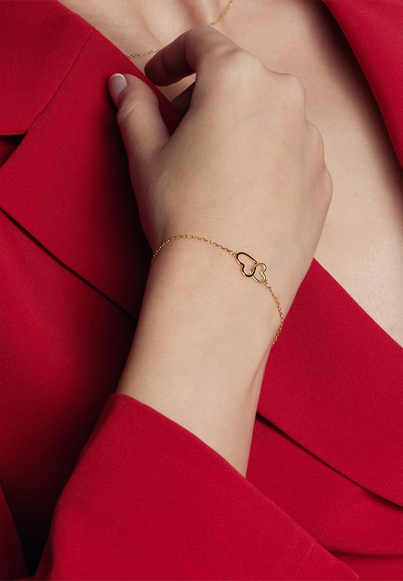 Złota bransoletka w kształcie serca z cienkim łańcuszkiem, noszona na dłoni na tle czerwonego materiału. Prosty design z polerowanym wykończeniem.