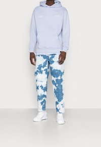 Felpa azzurro chiaro con tasca a marsupio e logo bianco. Pantaloni in denim strappati con un motivo di nuvole blu e bianche. Scarpe da ginnastica bianche.