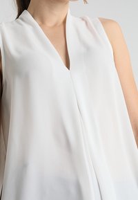 Blusa bianca senza maniche in tessuto leggero, con scollatura a V e design fluido. La texture appare liscia con una leggera lucentezza.