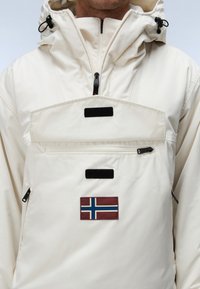 Mann trägt eine elfenbeinfarbene Kapuzenjacke mit Frontreißverschlusstasche und einem kleinen norwegischen Flaggenaufnäher auf der Brust.