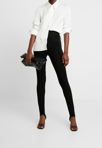 Vit skjorta med krage, slips, svarta ribbade leggings och en svart kuvertväska. Svarta högklackade sandaler. Enkel, strukturerad design.