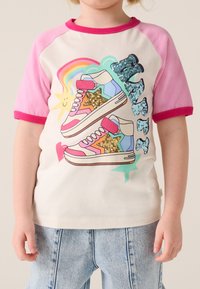 Īsām piedurknēm krekls ar rozā piedurknēm un baltu korpusu, uz kura attēloti krāsaini sporta apavi ar spīdīgām detaļām un rotaļīgiem dizainiem.