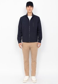 Veste de bombardier navy avec fermeture éclair à l'avant et poches latérales, portée sur une chemise blanche, associée à un pantalon beige et des baskets blanches.