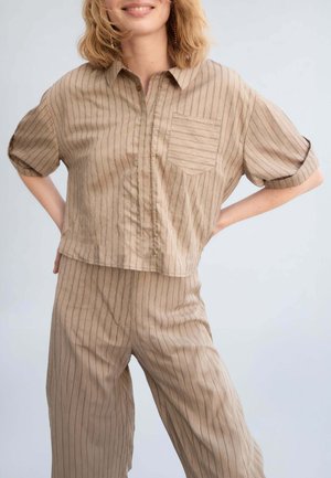 Donna che indossa una camicia beige a righe sottili a maniche corte con bottoni e pantaloni vita alta abbinati, in piedi con le mani sui fianchi.