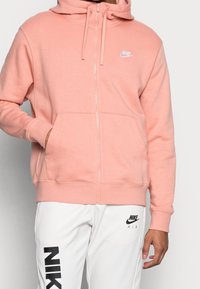 Ljusrosa zip-up hoodie i mjukt tyg, med en framficka och vitt Nike-logotyp på bröstet. Matchas med vita byxor.