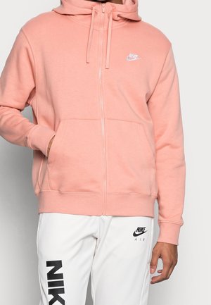 Sudadera con cremallera de color rosa claro, hecha de un tejido suave, que presenta un bolsillo frontal y el logo blanco de Nike en el pecho. Combinada con pantalones blancos.