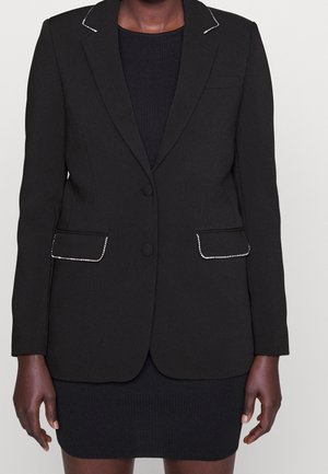 Schwarzer taillierter Blazer mit silbernen Perlenverzierungen am Kragen und an den Taschenklappen, getragen über einem schwarzen, gerippten Kleid.