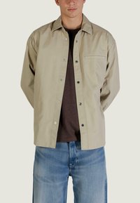 Camicia beige chiaro con chiusura a bottoni, taglio dritto, taschino sul petto e bottoni a pressione argentati, indossata sopra una T-shirt scura.