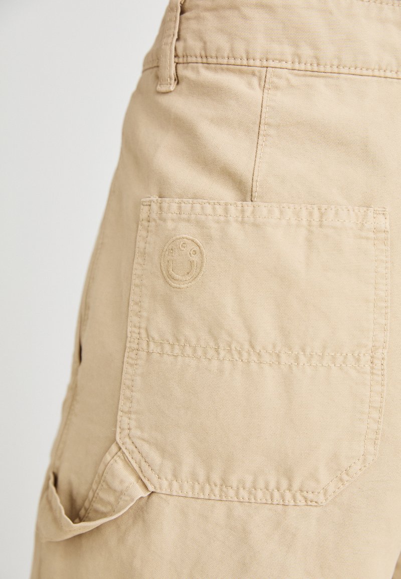 Pantaloni corti in tela beige con una grande tasca applicata, con logo ricamato di faccina sorridente. Vestibilità dritta e dettagli di cucitura rinforzata.