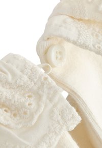 Vêtement couleur crème avec des motifs floraux brodés, doté d'un bouton décoratif et d'un tissu doux et texturé.