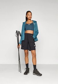 Teal zip-up hoodie over a dark sports bra, paired with black cargo shorts. Black hiking boots, trekking poles in hand, neutral backdrop.